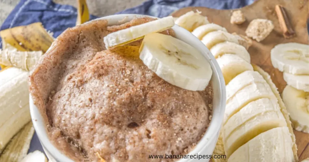 Close-up of a moist and fluffy banana bread mug cake topped with chocolate chips, served in a white mug on a wooden table, with ripe bananas in the background.
